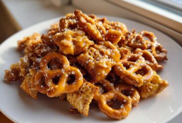 A pile of crunchy pretzels and cereal coated in shiny toffee glaze, sprinkled with coarse sugar, part of the toffee chex mix recipe.