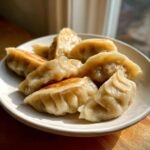 A plate of golden, pan-fried Traditional Chinese Dumplings with pleated edges, catching the sunlight.