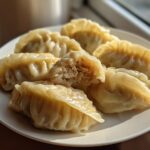 Close-up of several perfectly pleated Traditional Chinese Dumplings served on a white plate, one is broken open showing the filling.