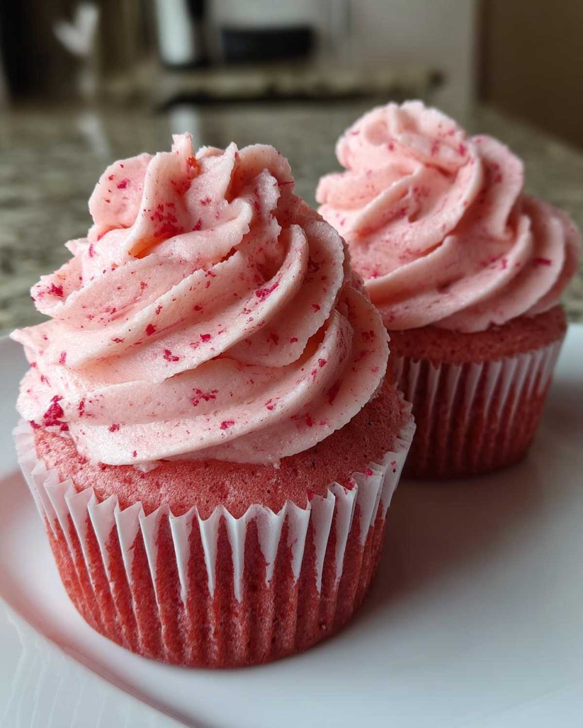 Close-up of two perfectly frosted strawberry cupcakes with pink swirl frosting flecked with red pieces.