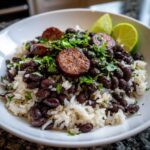 A close-up of a bowl of seasoned black beans and rice topped with sliced sausage and fresh cilantro, served with lime wedges.