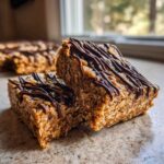 Two thick, chewy homemade protein bars stacked slightly, drizzled generously with dark chocolate.