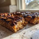 Four homemade protein bars topped with melted chocolate drizzle sitting on a marble counter near a window.