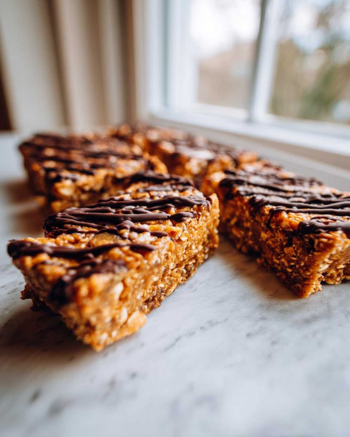 Close-up of several homemade protein bars drizzled with dark chocolate, resting on a white marble surface.