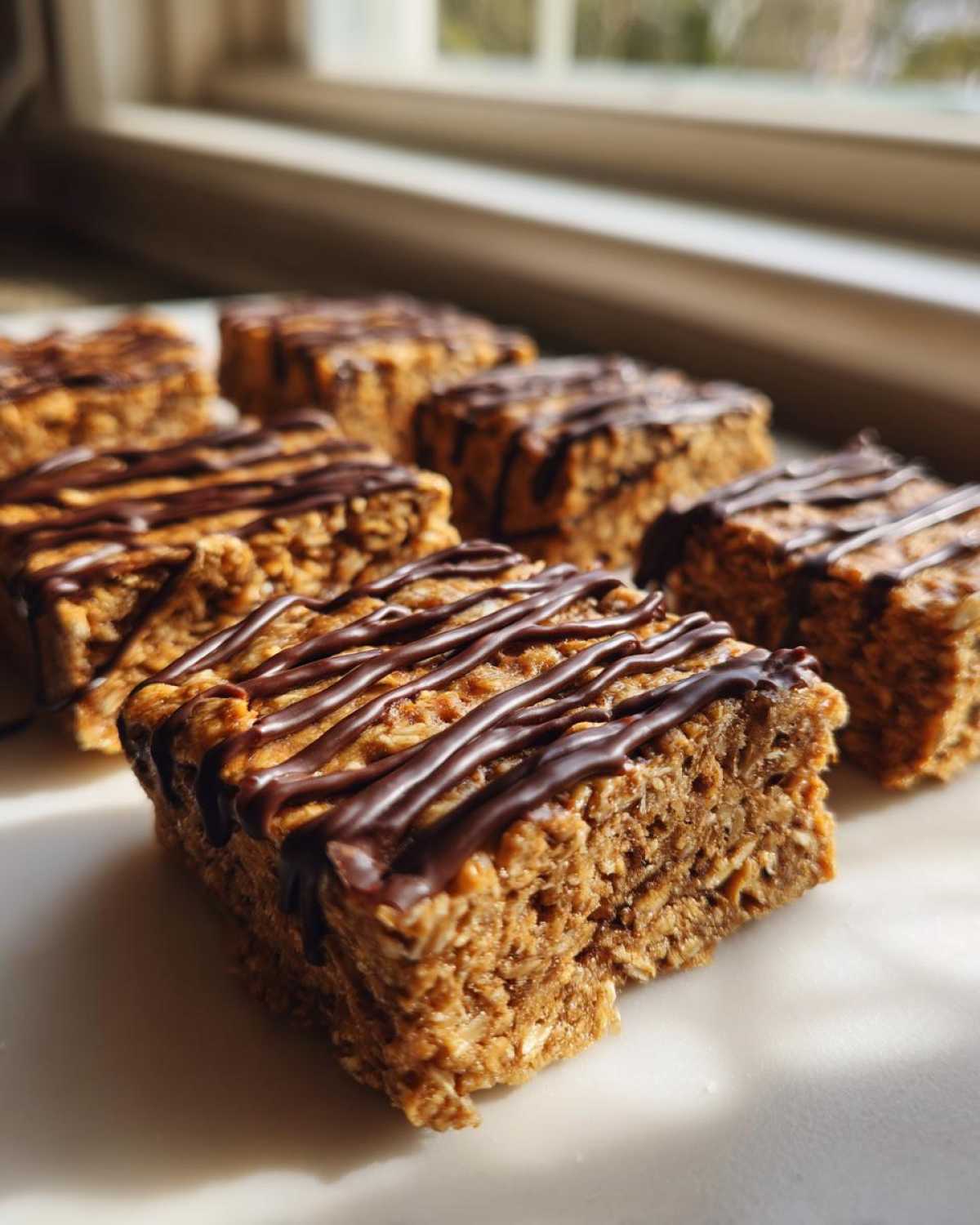 Close-up of several homemade protein bars drizzled with dark chocolate, set near a window.