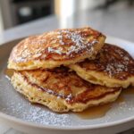 A stack of three fluffy almond flour pancakes drizzled with syrup and dusted heavily with powdered sugar.