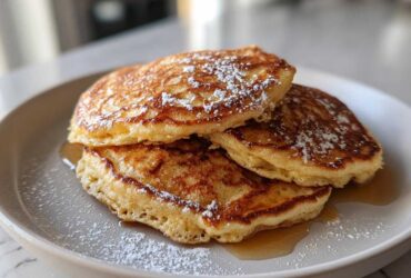A stack of three fluffy almond flour pancakes drizzled with syrup and dusted heavily with powdered sugar.