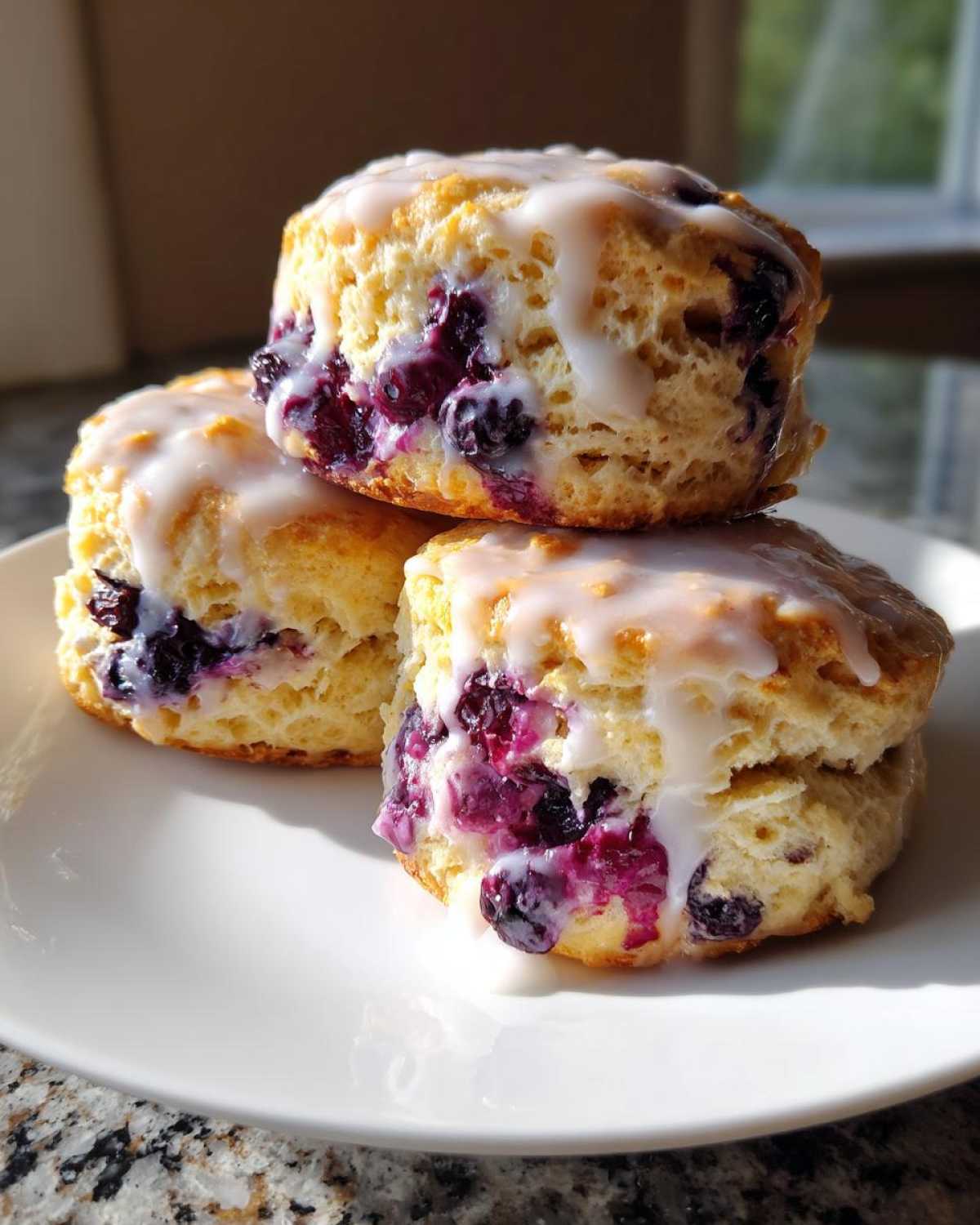 Three fluffy blueberry scones stacked on a white plate, topped with a sweet white glaze and bursting with purple blueberries.