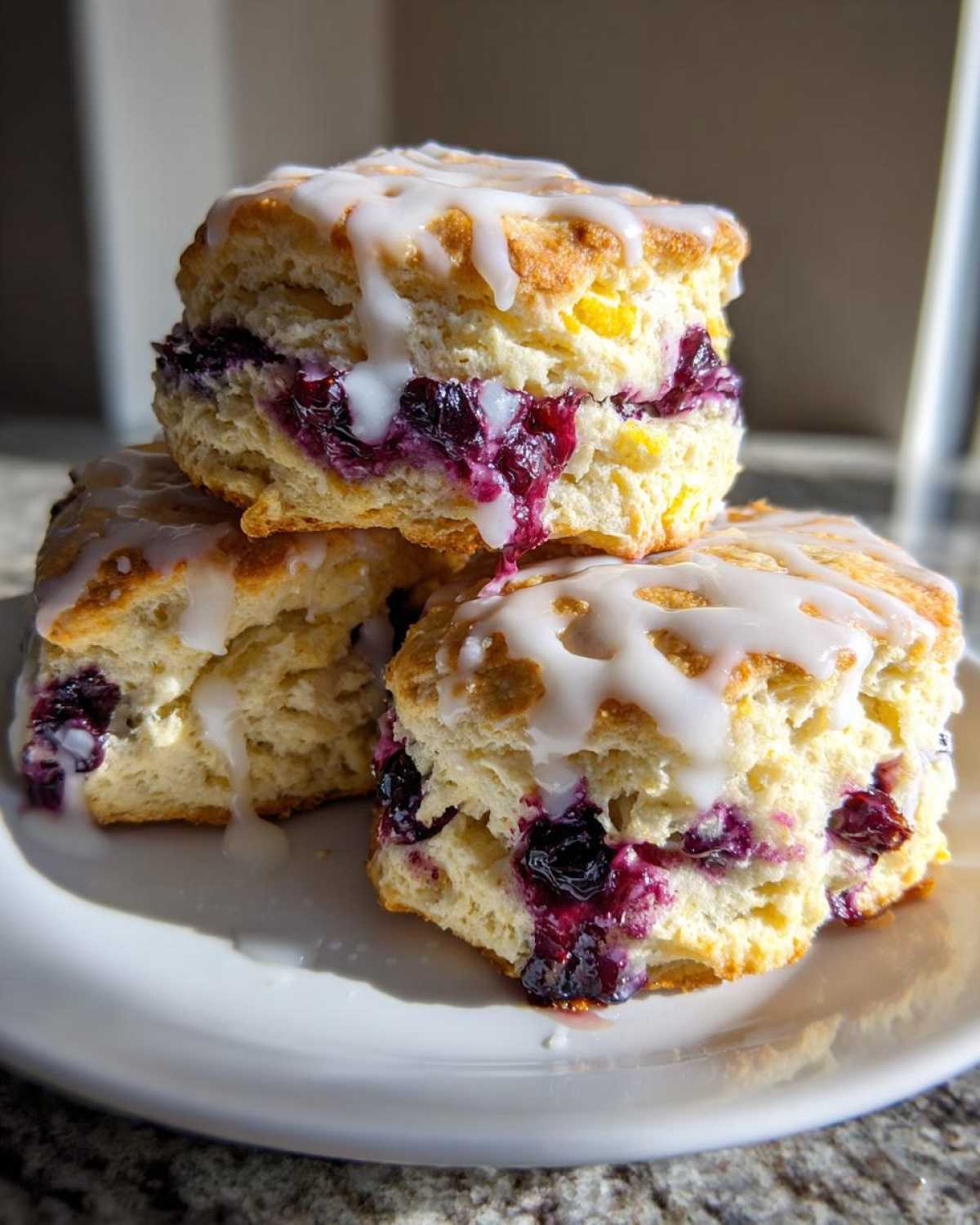 Three stacked, flaky blueberry scones topped with a thick white glaze, showing juicy, purple blueberry filling.