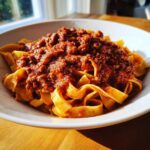 A white bowl filled with wide egg noodles topped generously with a thick, rich bolognese sauce.