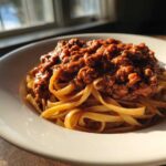 A close-up of fettuccine pasta topped generously with rich bolognese sauce, backlit by a window.