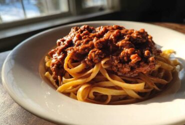 A close-up of fettuccine pasta topped generously with rich bolognese sauce, backlit by a window.