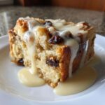 A close-up of a slice of moist bread pudding loaded with raisins, drizzled generously with vanilla cream sauce.