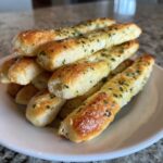 A stack of freshly baked, golden brown breadsticks seasoned with garlic and parsley served on a white plate.