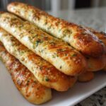 A close-up stack of golden, freshly baked breadsticks topped with garlic butter and parsley.