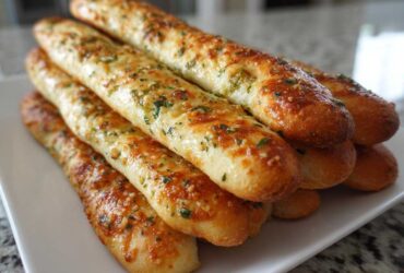 A close-up stack of golden, freshly baked breadsticks topped with garlic butter and parsley.