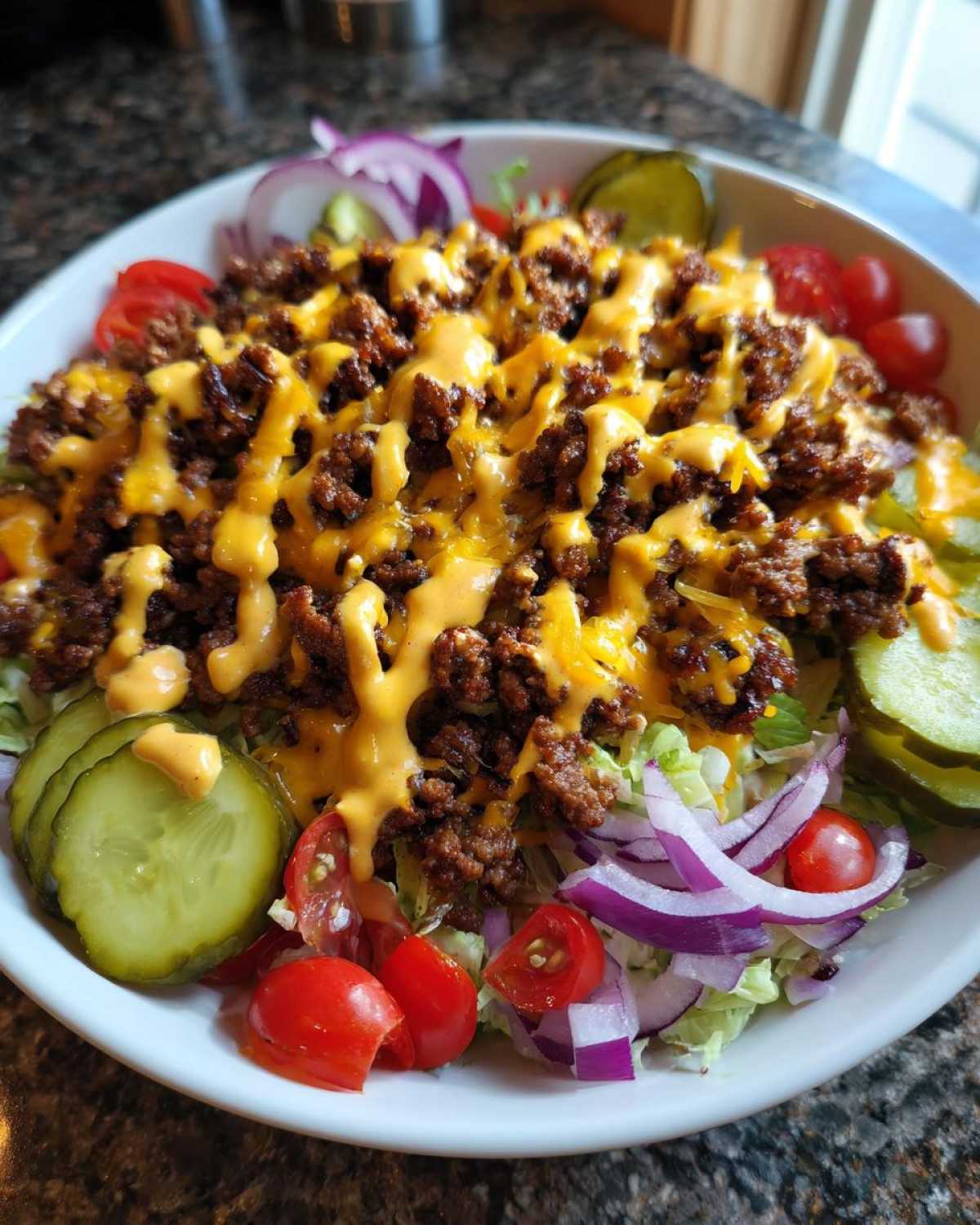 A vibrant cheeseburger bowl featuring seasoned ground beef, melted cheese, and special sauce over lettuce, garnished with pickles, tomatoes, and red onion.