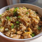 A close-up of a white bowl filled with delicious chicken fried rice, featuring visible pieces of chicken, scrambled egg, and fresh green onions.