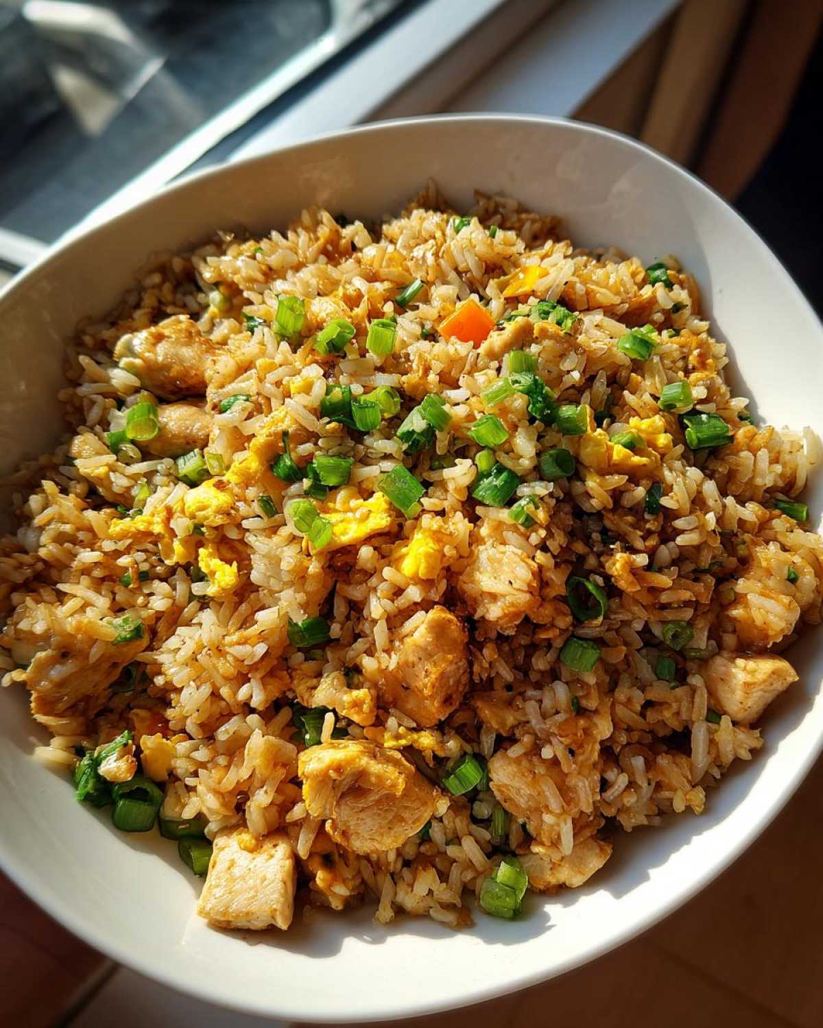 A close-up overhead view of a white bowl filled with amazing chicken fried rice, featuring chunks of chicken, scrambled egg, and green onions.