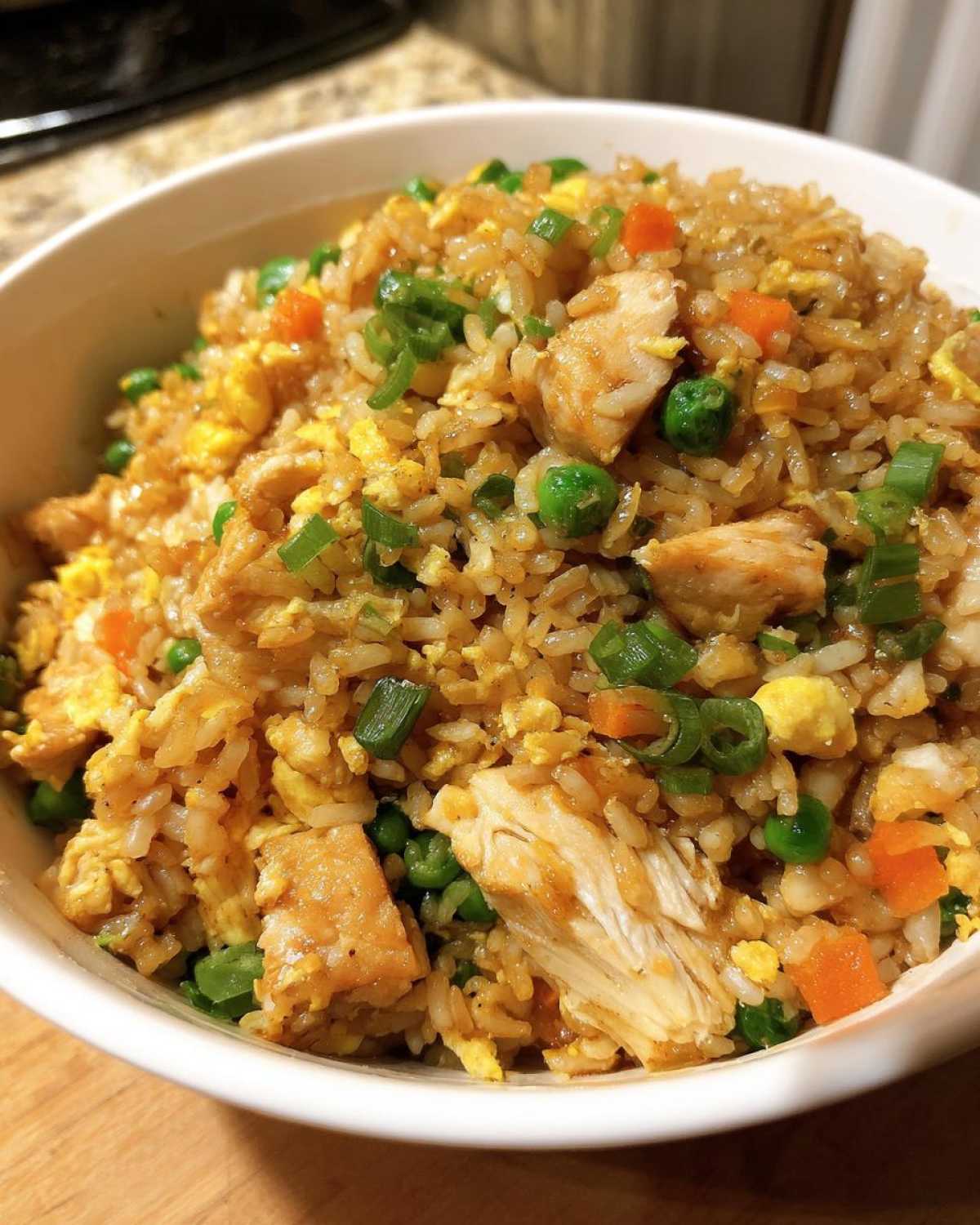 Close-up of a white bowl filled with amazing chicken fried rice, featuring chunks of chicken, peas, carrots, egg, and green onions.