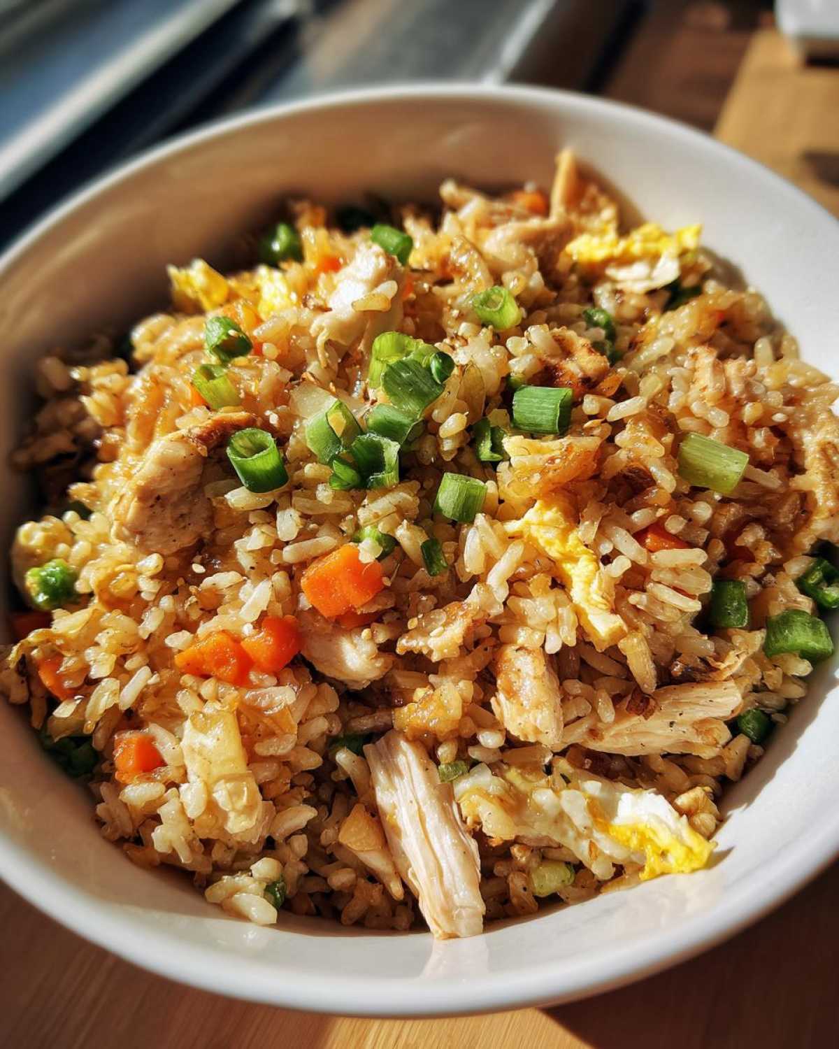 A close-up of a white bowl filled with amazing chicken fried rice, featuring shredded chicken, carrots, egg, and green onions.