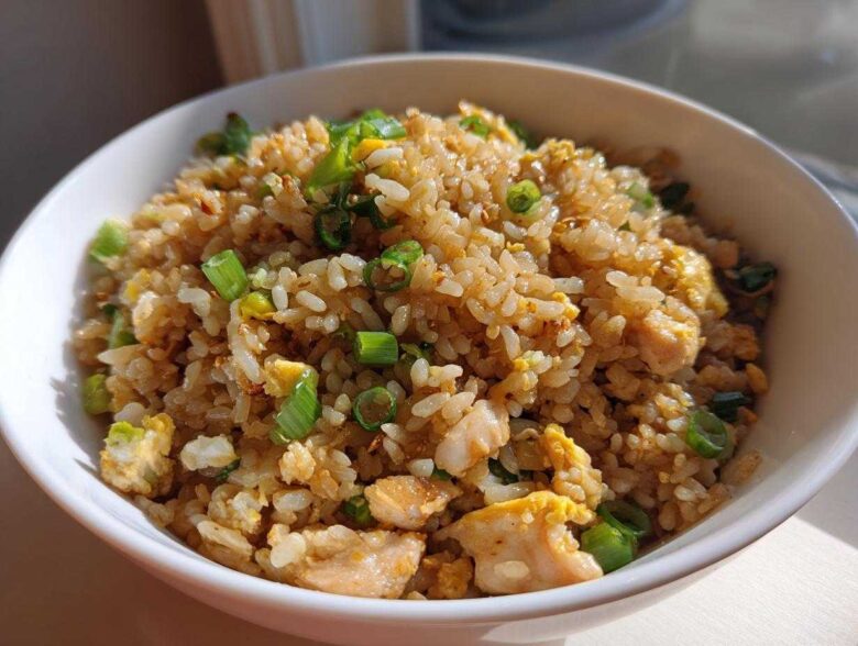 A close-up of a white bowl filled with delicious chicken fried rice, featuring visible pieces of chicken, scrambled egg, and fresh green onions.