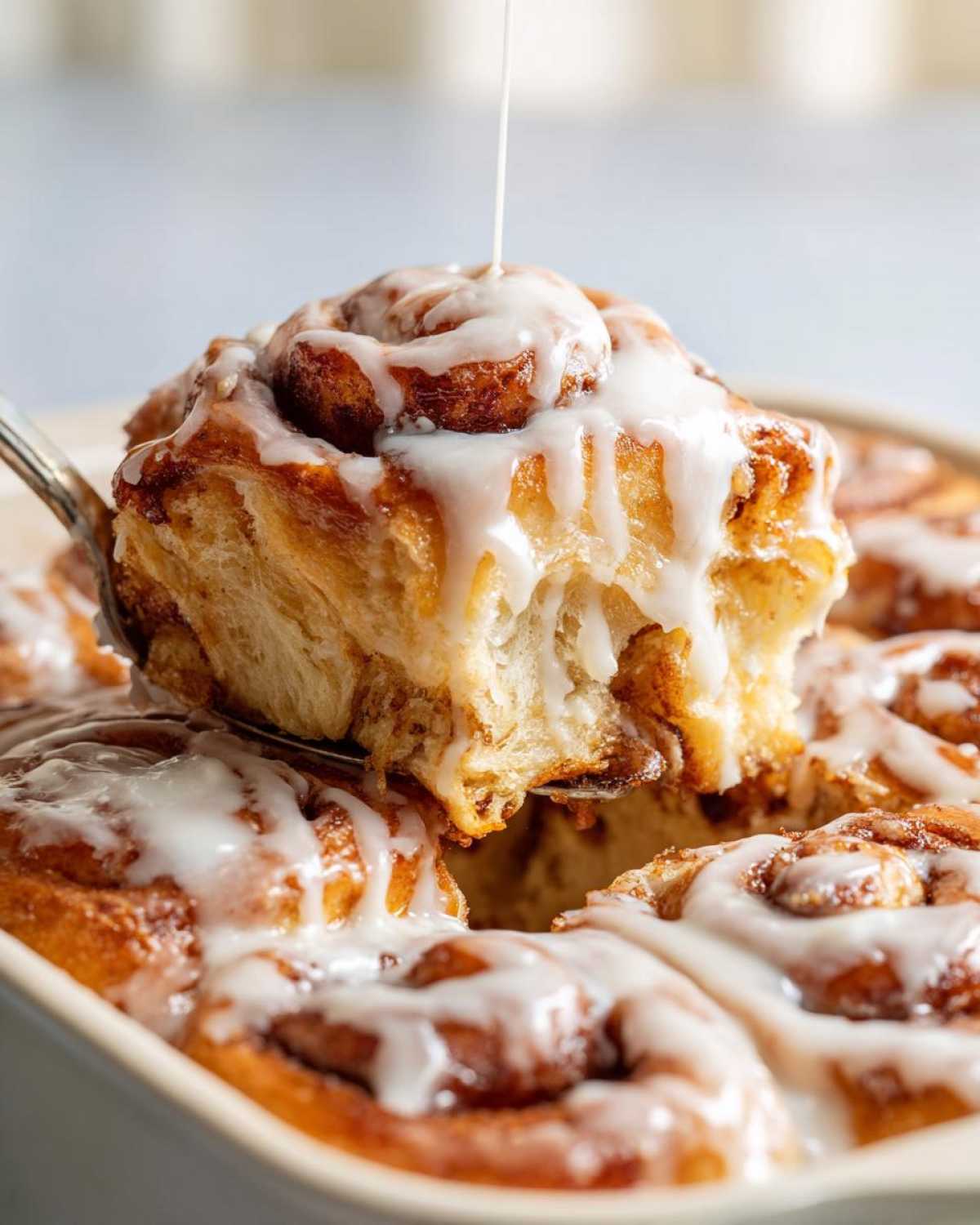 A serving spoon lifts a gooey, iced piece of Cinnamon roll casserole from the baking dish.