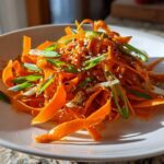 Close-up of a vibrant raw carrot salad made with shaved carrots, topped with sesame seeds and sliced green onions.