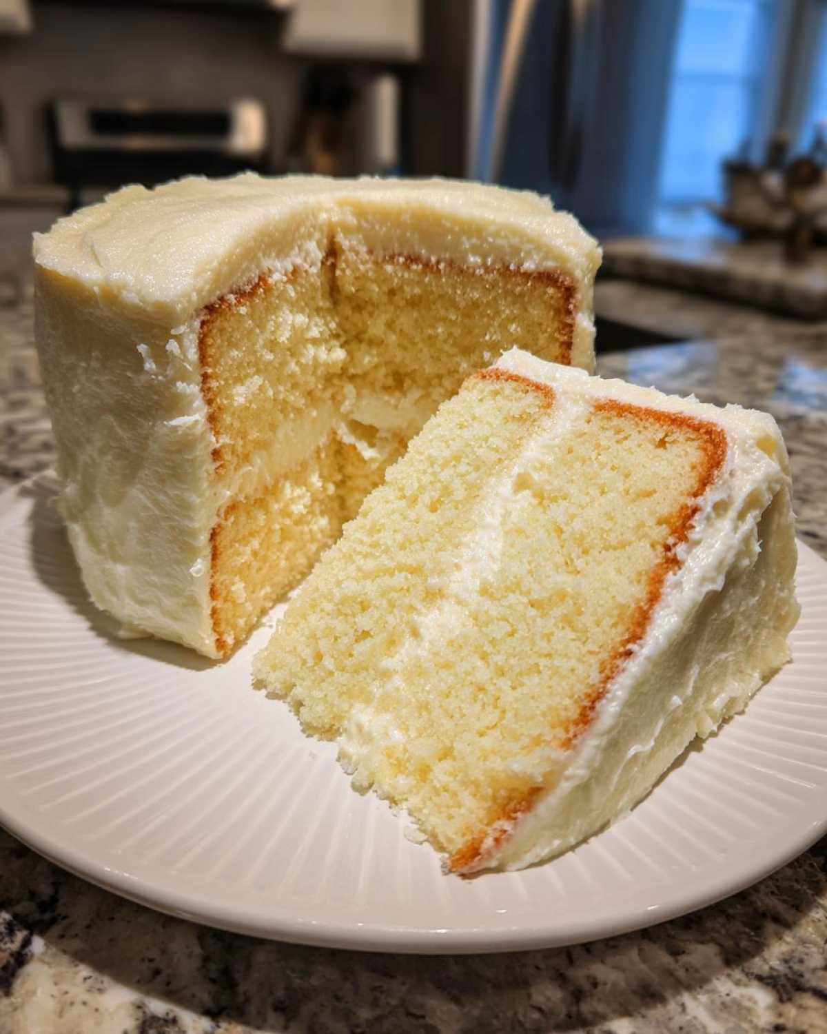 A slice of moist vanilla cake recipe with thick white frosting, sitting next to the rest of the cake on a white plate.