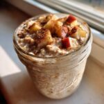 Close-up of apple cinnamon overnight oats in a glass jar, topped with diced apples and cinnamon.