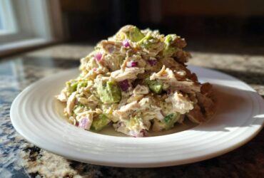 A serving of creamy avocado chicken salad mixed with chunks of avocado and red onion on a white plate.
