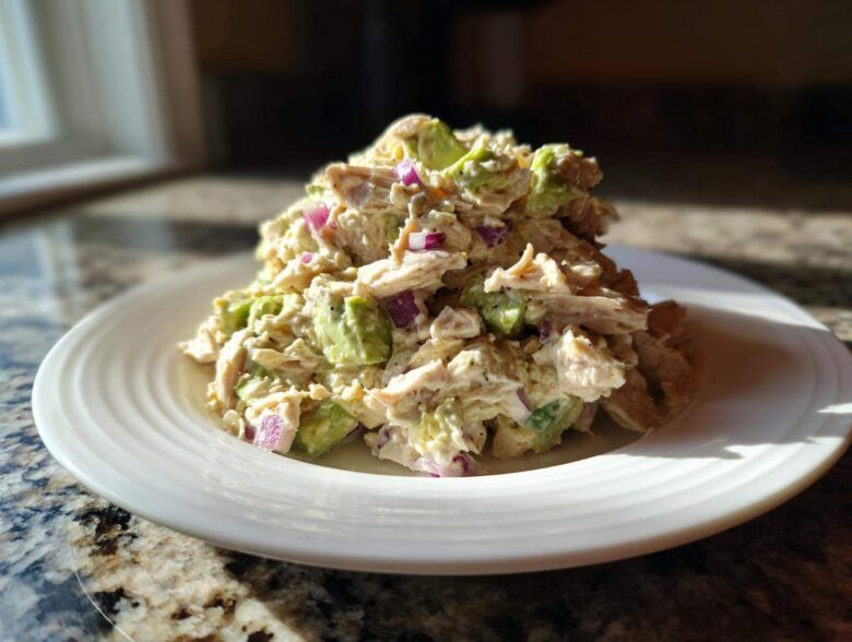 A serving of creamy avocado chicken salad mixed with chunks of avocado and red onion on a white plate.