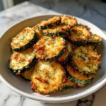 A close-up of golden brown, crispy baked parmesan zucchini slices piled high in a light-colored bowl.