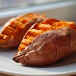 Two perfectly baked sweet potatoes, scored and fluffed, sitting on a white plate.