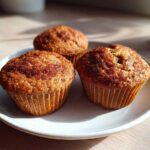 Three freshly baked banana protein muffins dusted with cinnamon sugar sitting on a white plate.