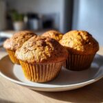 Close-up of four golden-brown banana protein muffins sitting on a light plate, highlighted by sunlight.