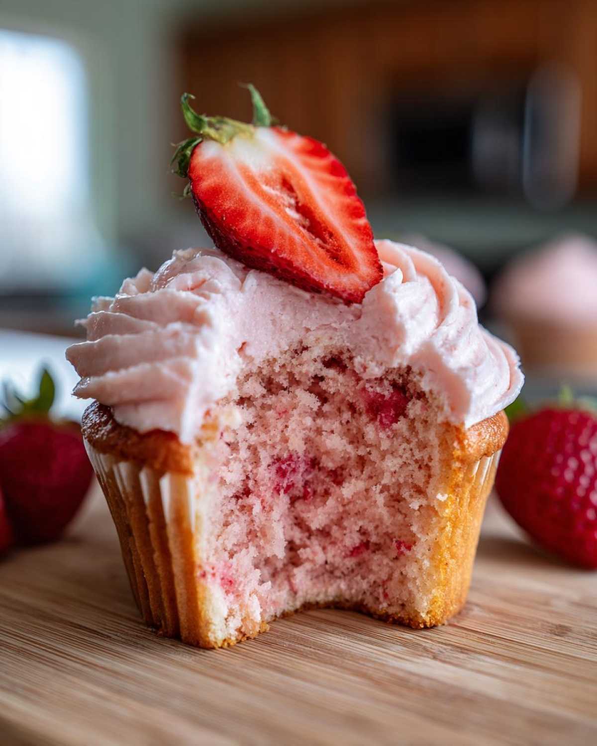 Close-up of a strawberry cupcake with a bite taken out, showing pink cake, frosting, and a fresh strawberry half on top.
