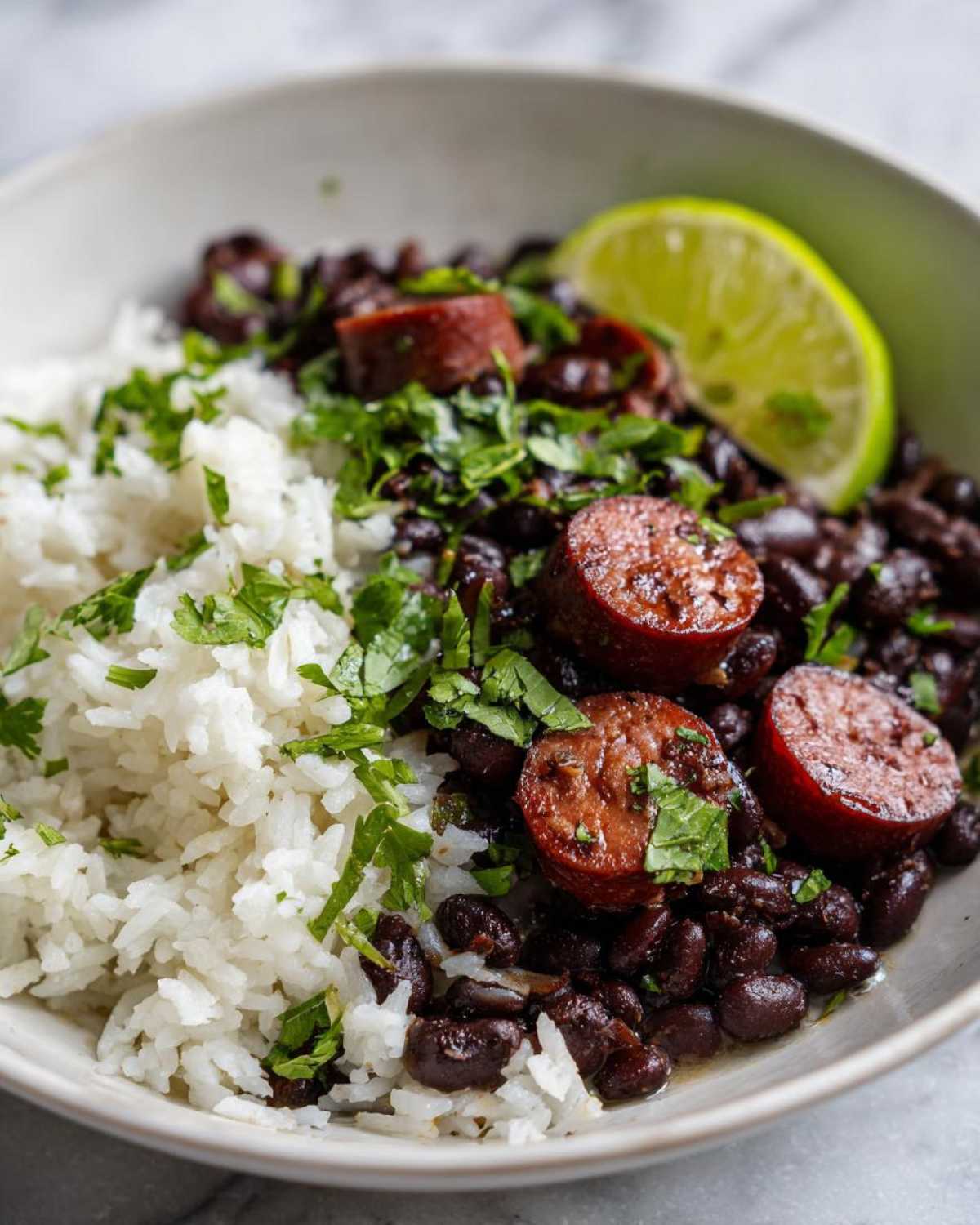 Close-up of a bowl of flavorful black beans and rice topped with sliced sausage, cilantro, and a lime wedge.