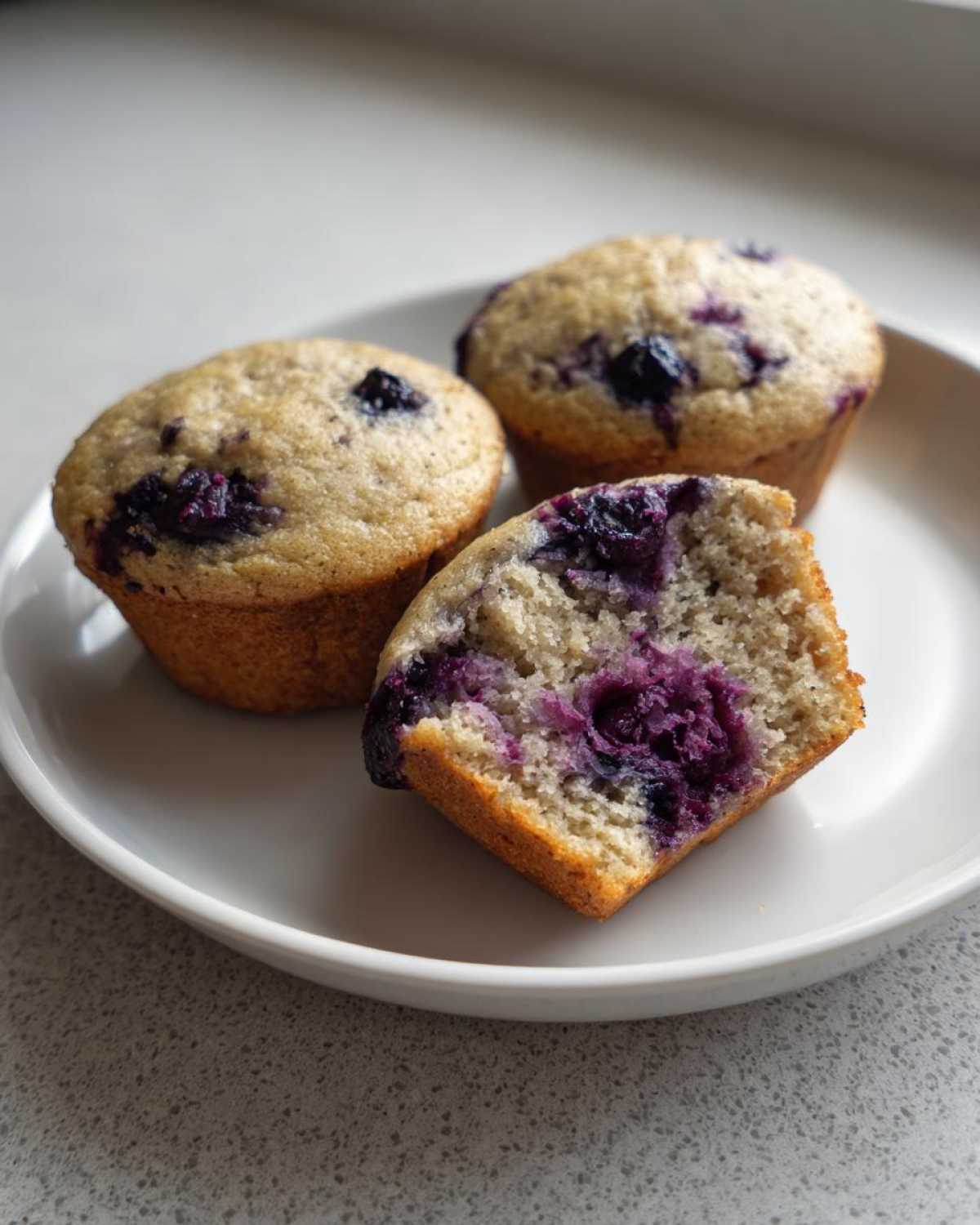 Three blueberry protein muffins on a white plate, one cut in half showing the moist interior.