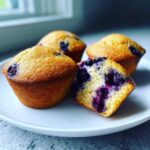 Four golden-brown blueberry protein muffins on a white plate, one cut in half showing the moist interior.