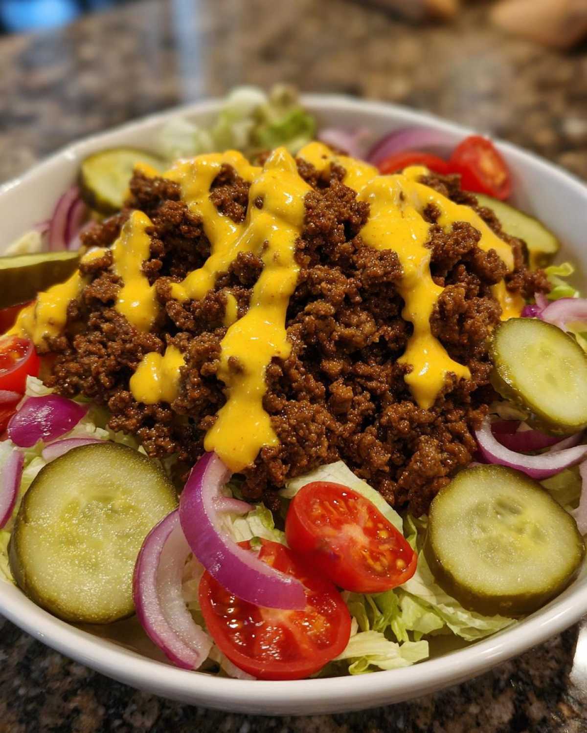 A close-up of an amazing cheeseburger bowl featuring seasoned ground beef, bright yellow cheese sauce, lettuce, pickles, red onion, and cherry tomatoes.