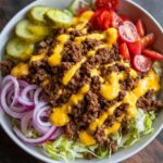 A vibrant cheeseburger bowl featuring seasoned ground beef, cheese sauce, pickles, tomatoes, and red onion over lettuce.
