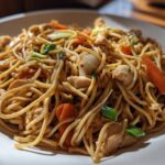 Close-up of a white plate piled high with saucy chicken chow mein recipe noodles, chicken pieces, and sliced carrots.
