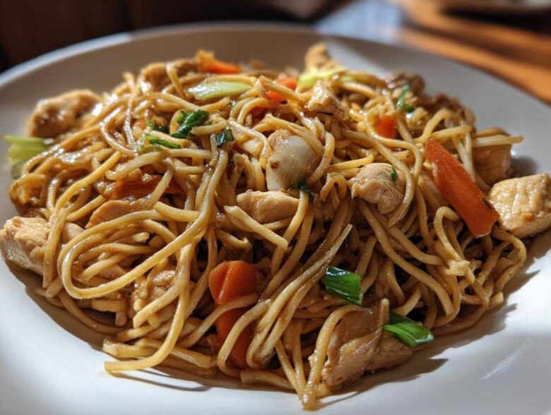Close-up of a white plate piled high with saucy chicken chow mein recipe noodles, chicken pieces, and sliced carrots.