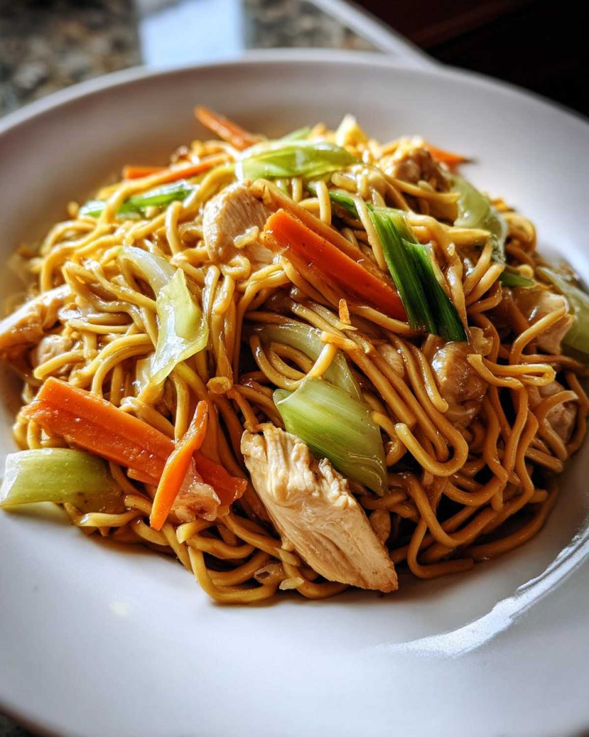 Close-up of a white bowl filled with savory chicken chow mein recipe, featuring noodles, sliced chicken, carrots, and bok choy.