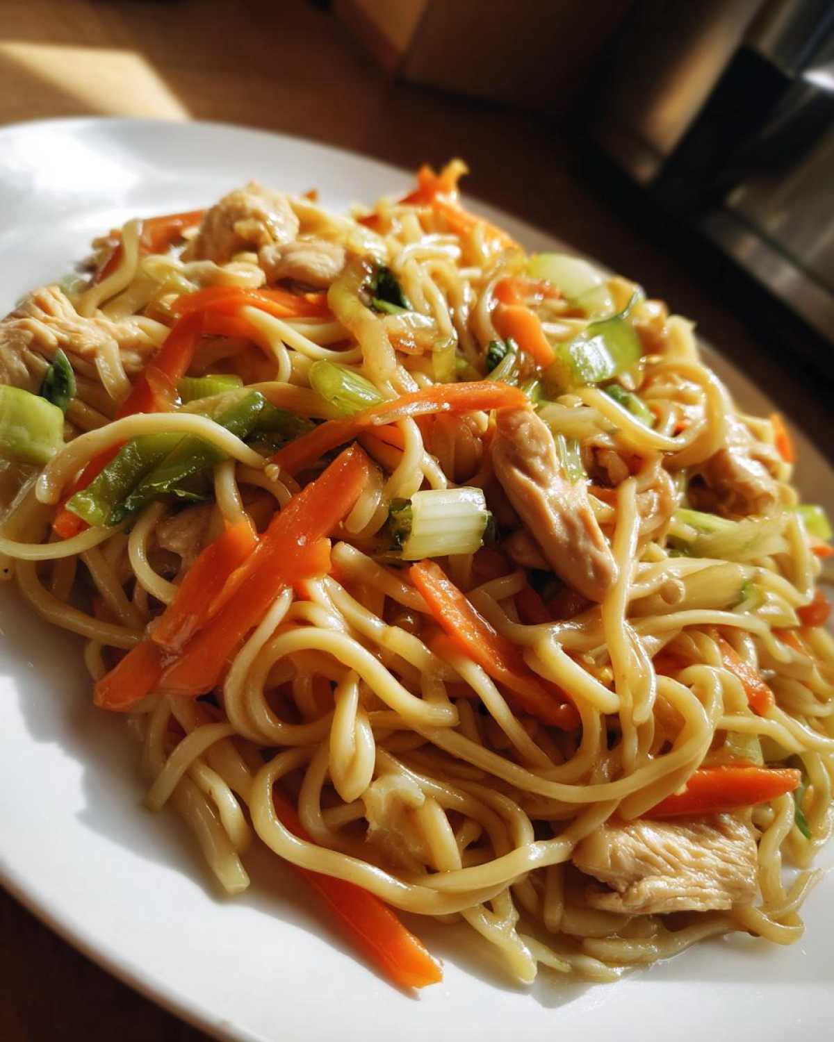 Close-up of a plate piled high with stir-fried chicken chow mein recipe, featuring noodles, sliced carrots, and green bok choy.