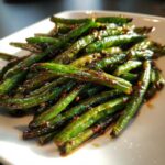 A close-up of glossy, stir-fried chinese green beans with garlic sauce piled on a white plate.