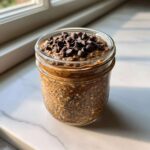 A glass jar filled with thick chocolate overnight oats speckled with chia seeds, topped generously with chocolate chips.