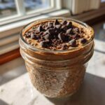 Close-up of chocolate overnight oats in a mason jar topped with chocolate chips, sitting by a sunny window.