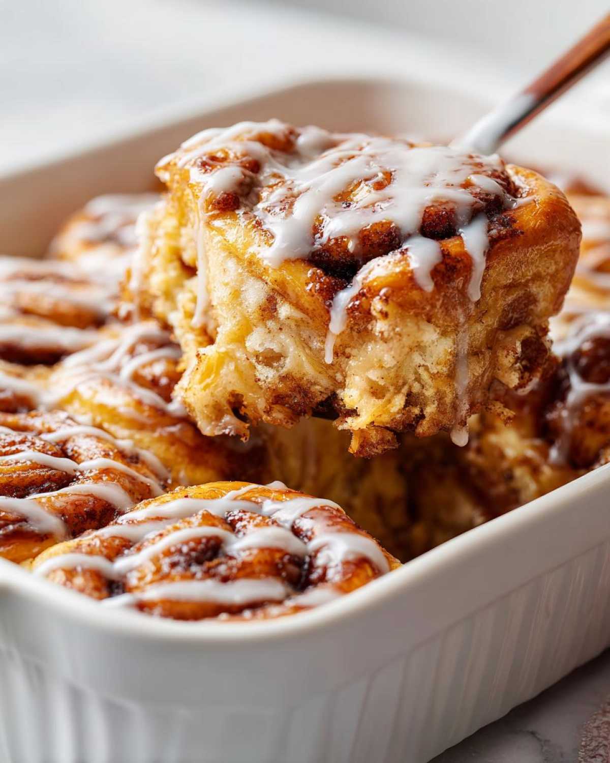 A serving spoon lifts a gooey piece of cinnamon roll casserole topped with white icing from a white baking dish.